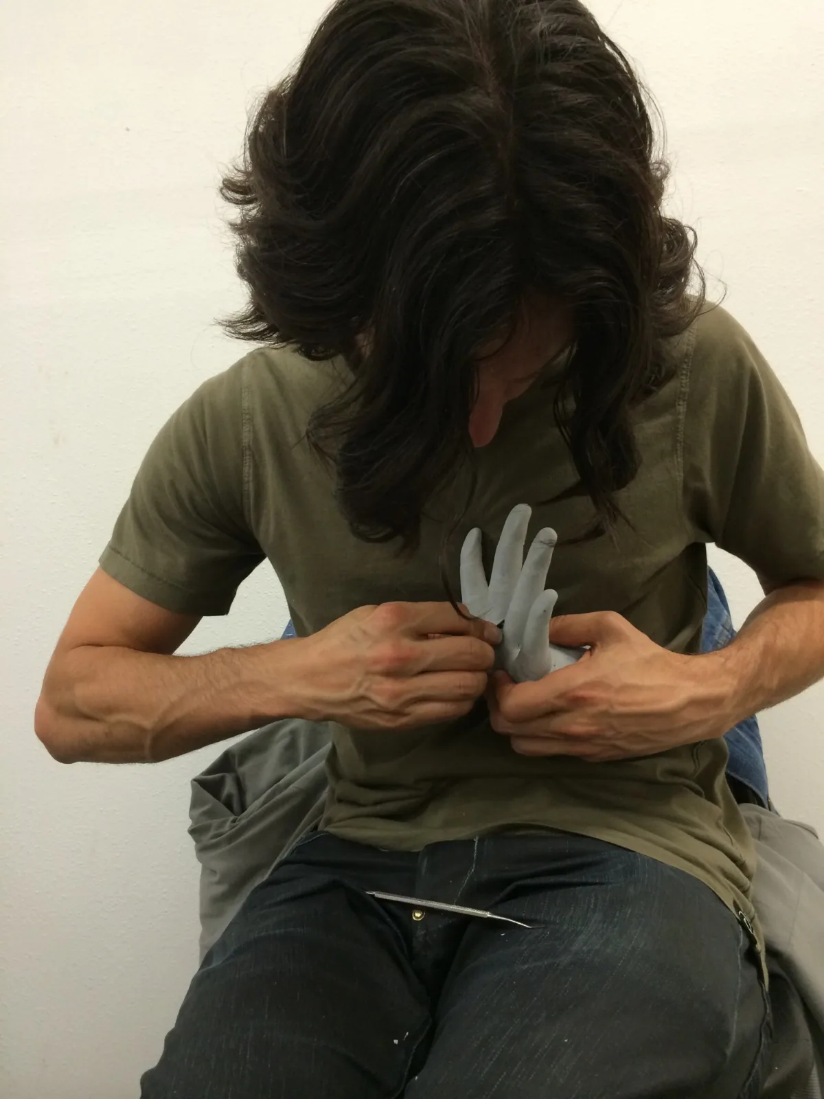 Cyrus, his hair falling over his face, working on a grey cast of a hand held to his chest, at DarkPlace