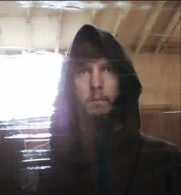 Cyrusrex in a hood, light flaring across the warehouse behind him