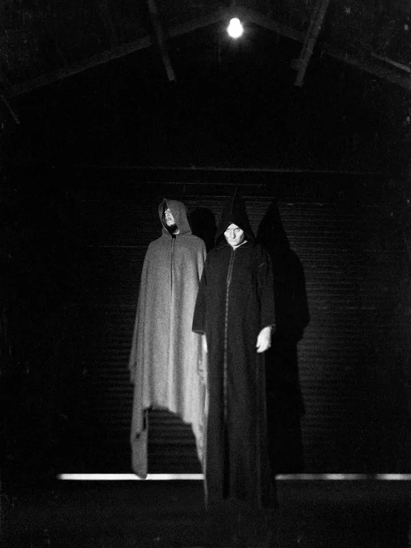 Two hooded figures in long robes, one white and one black, in a dark warehouse under a single bulb