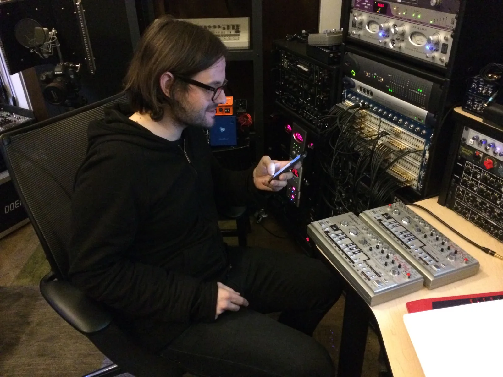 Alessandro Cortini in a studio chair, looking at his phone beside two Devil Fish TB-303s and racks of gear