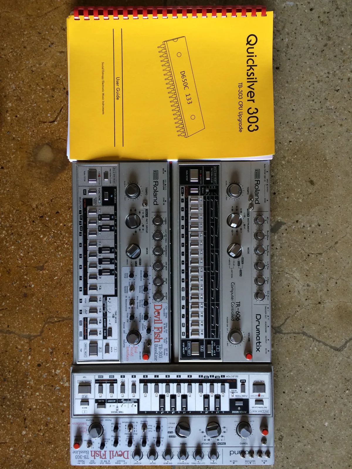 Two Devil Fish TB-303s and a TR-606 laid on a concrete floor under the yellow Quicksilver 303 user guide