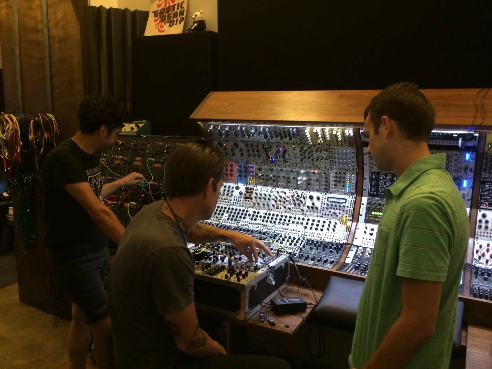 Three people at the modular systems: one patching a black system, one seated patching a case on the desk, one in a green shirt watching