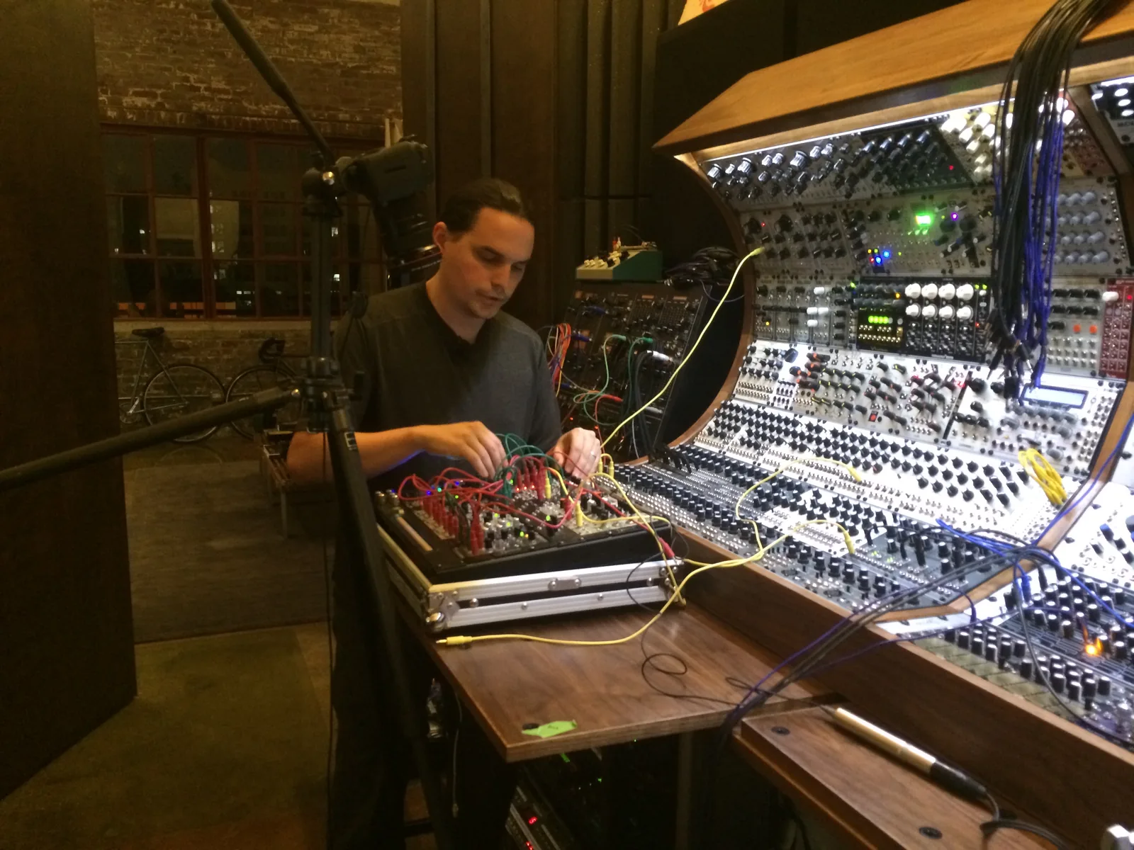A man at the curved modular console patching a case of modules, a camera on a tripod beside him