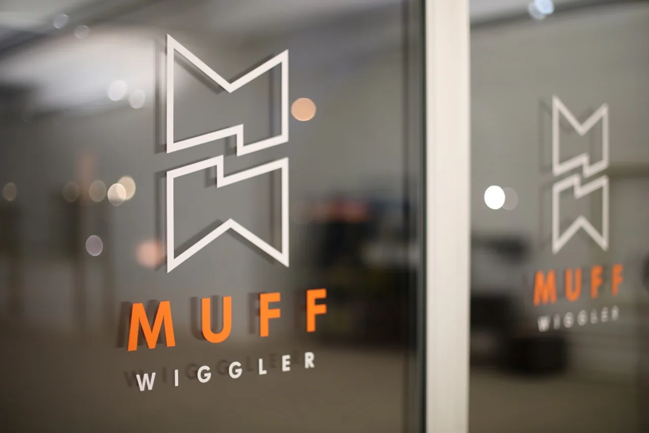 The Muff Wiggler logo in white and orange on the glass door of the Muffwiggler Store