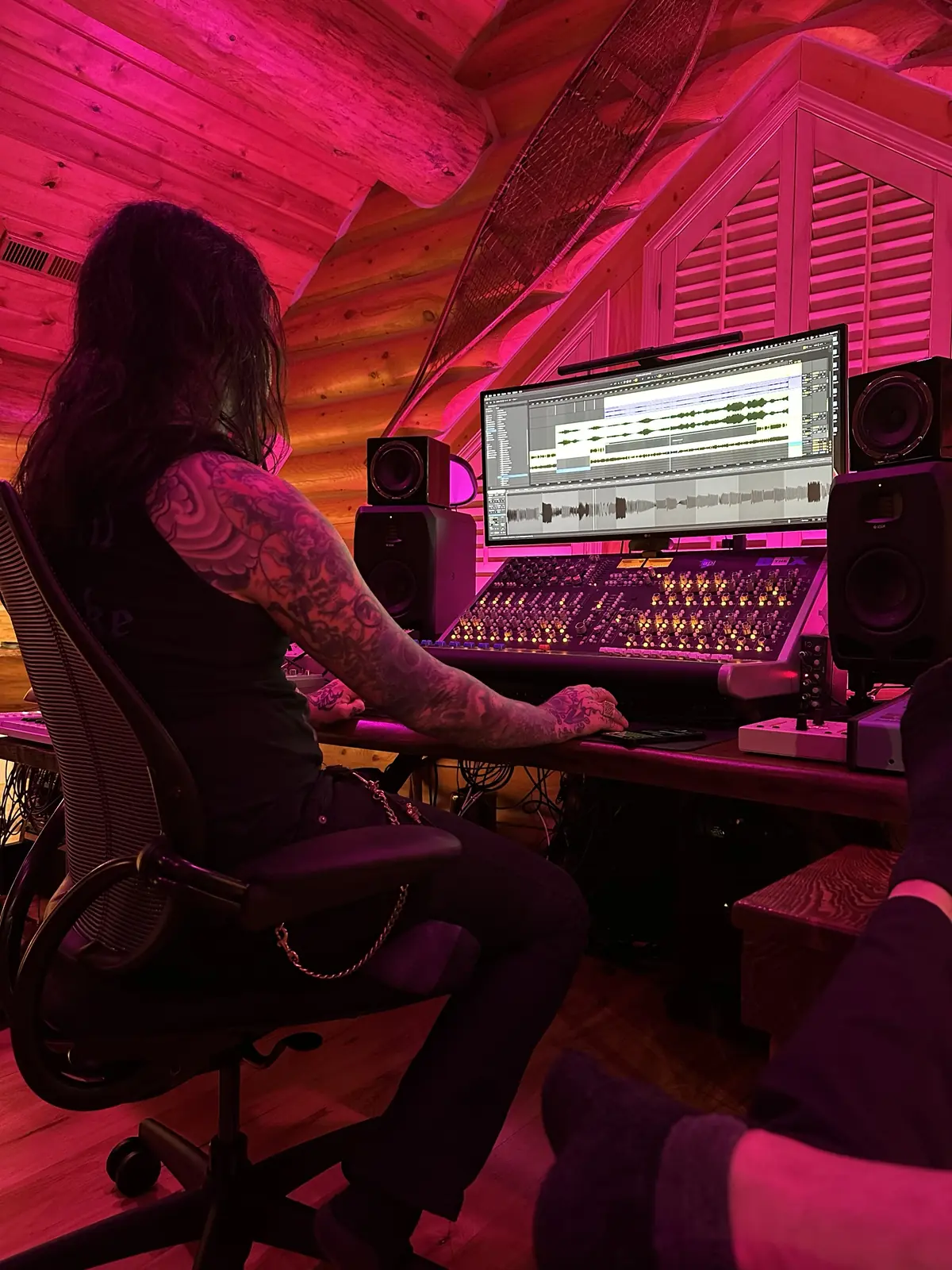A long-haired, tattooed musician seen from behind at a large mixing console under pink light, waveforms on a curved screen