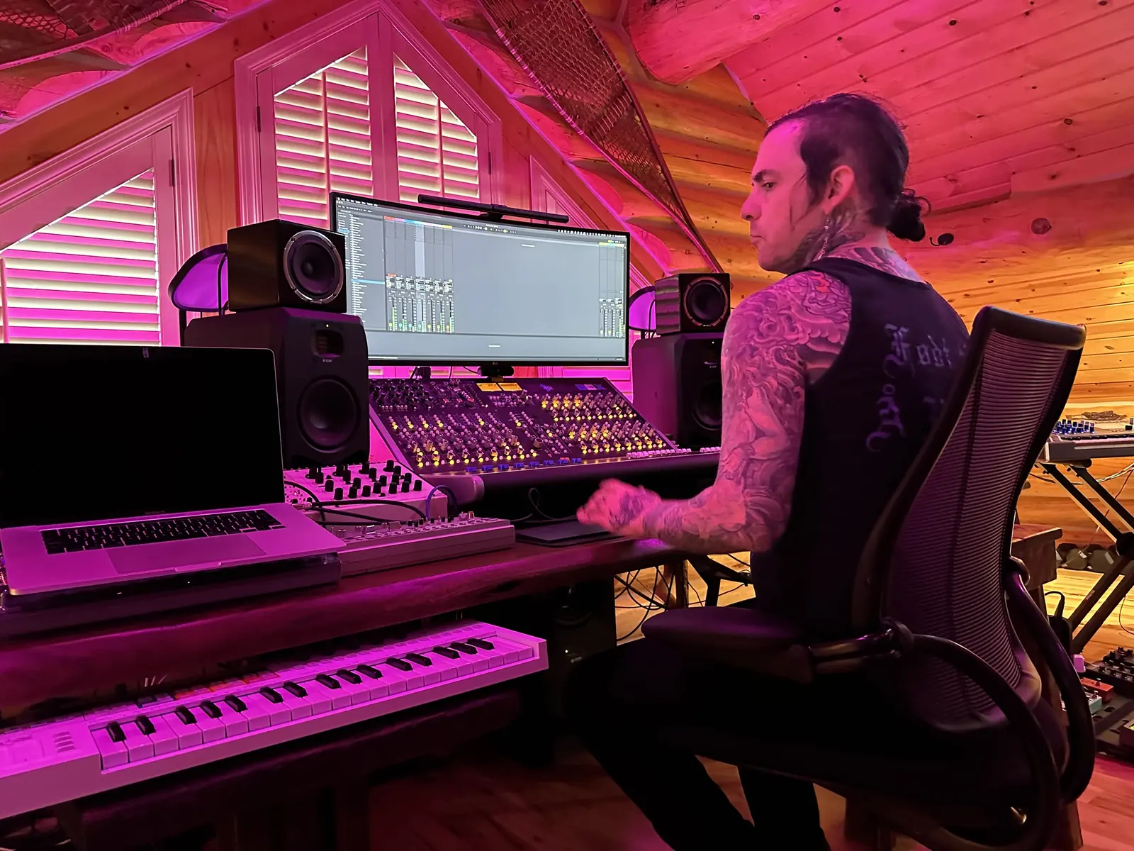 A tattooed musician at the mixing console beside a laptop and a keyboard, in the pink-lit log cabin studio