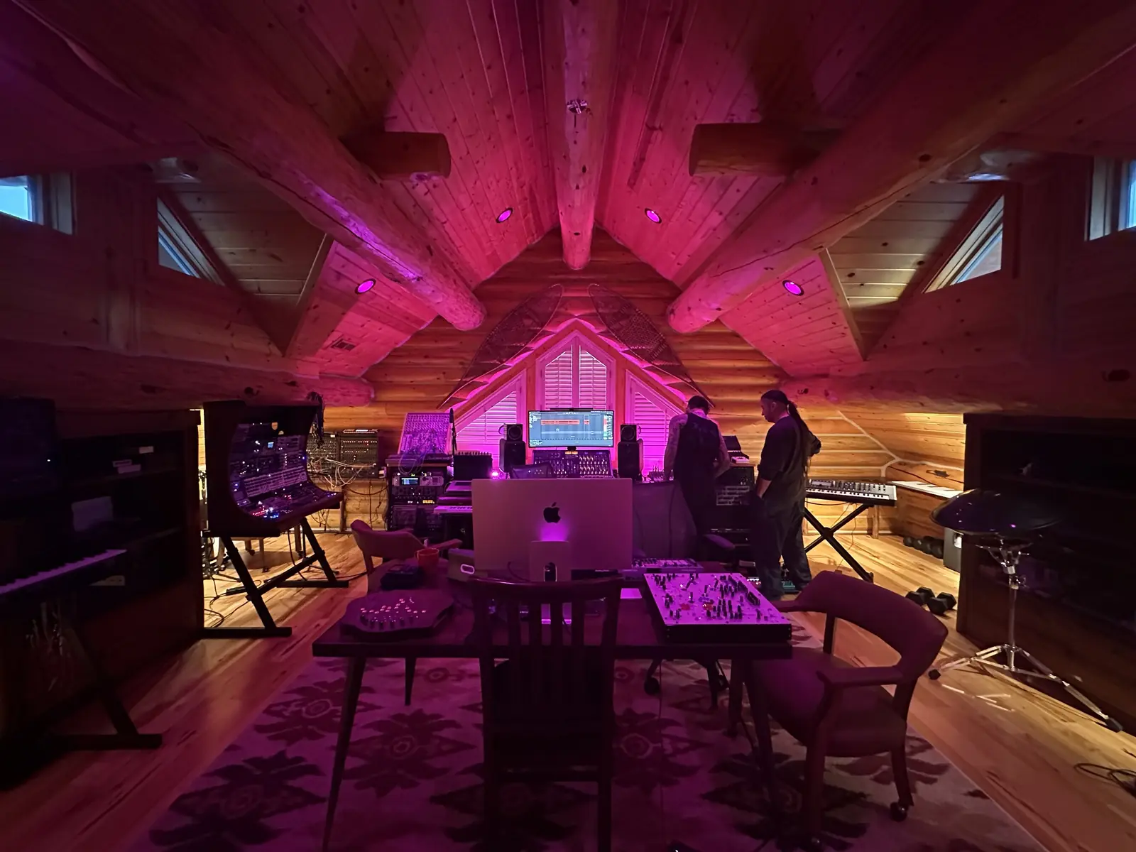 Wide view of the log cabin studio under pink light: modular cases, keyboards and the console, two people at the desk