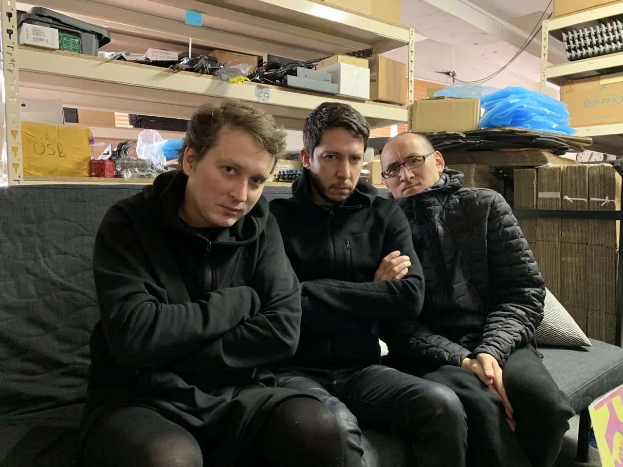 Three men on a couch at the Black Corporation studio in Tokyo, among them Roman and Richard Devine, shelves of parts behind
