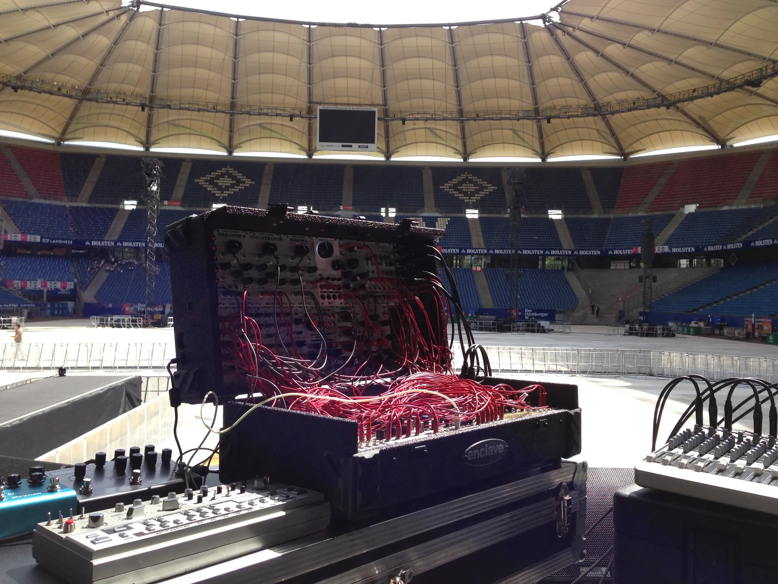 The suitcase modular, patched in red, with a TB-303 and effects pedals in front of it on a stadium stage, the stands empty
