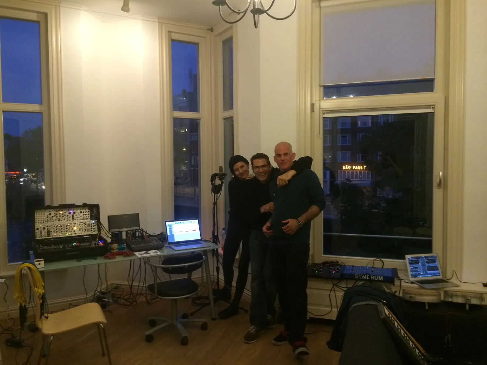 Three friends arm in arm in a tall-windowed room set up for recording, a modular case and laptops on the tables