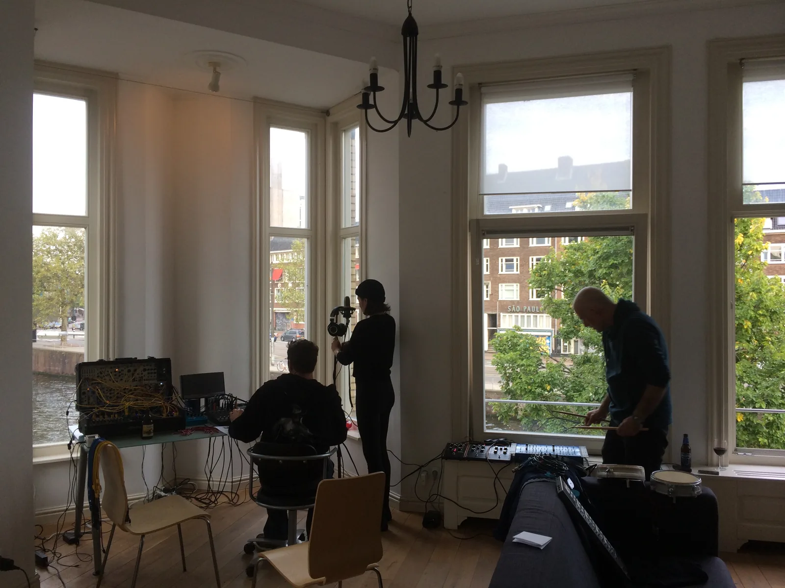 Recording in a tall-windowed room over a canal: one at the modular and a laptop, one holding a microphone, Ryan Moore at a drum