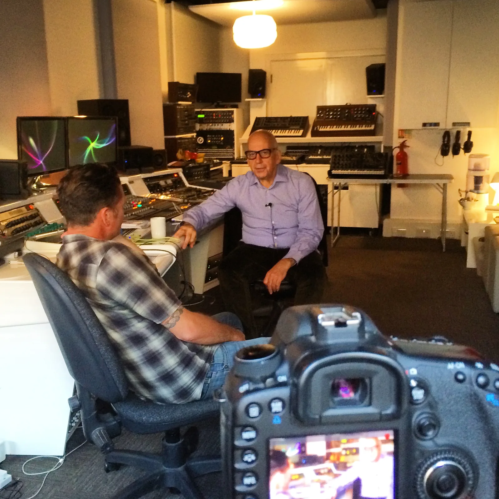 Daniel Miller talking with Douglas J. McCarthy in a studio lined with synthesizers, a camera in the foreground